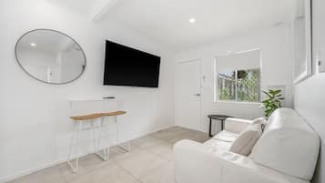 Un salón moderno con un sofá blanco, un televisor montado en la pared, un espejo redondo y una pequeña mesa con taburetes.