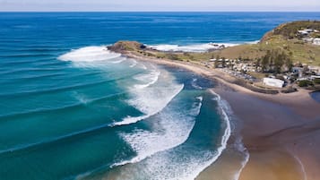 An aerial view of a coastal area with a beach, waves, and a small settlement with buildings.