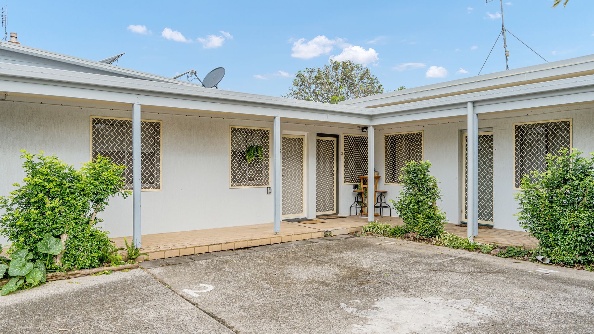 A two-story residential building with a covered patio area, satellite dishes, and a small garden.