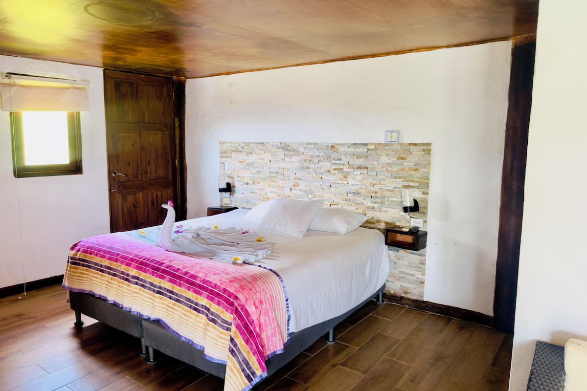 A bedroom with a bed, wooden floor, a stone headboard, a wooden door, and a window with curtains.