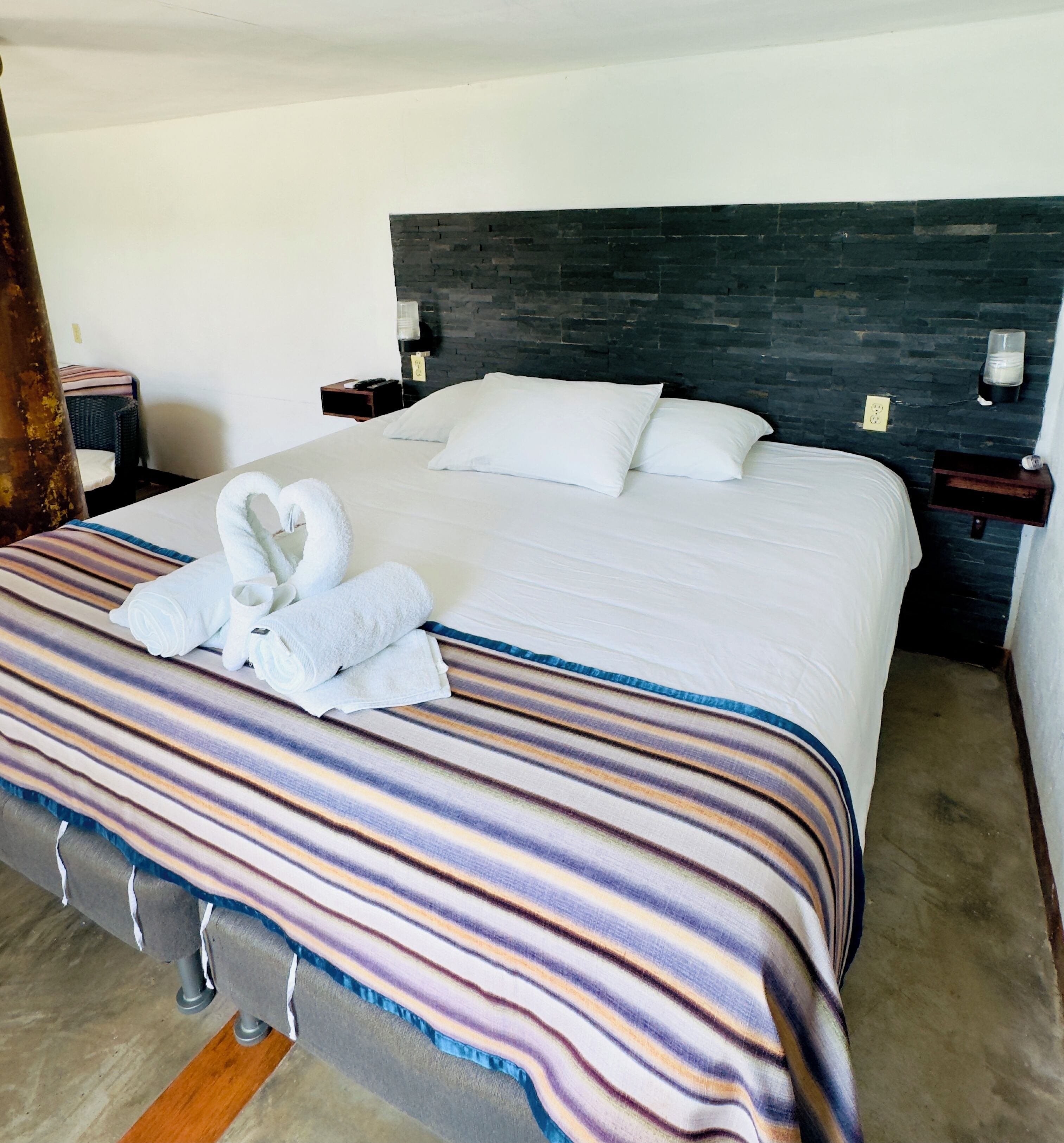 Uma cama bem arrumada com lençóis brancos e manta listrada, uma cabeceira de pedra escura e uma mesinha de cabeceira com abajur.