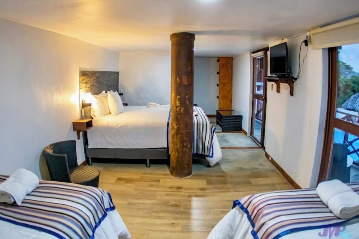 A hotel room with a large bed, two smaller beds, a wooden pillar, a TV, and a view of the outdoors.