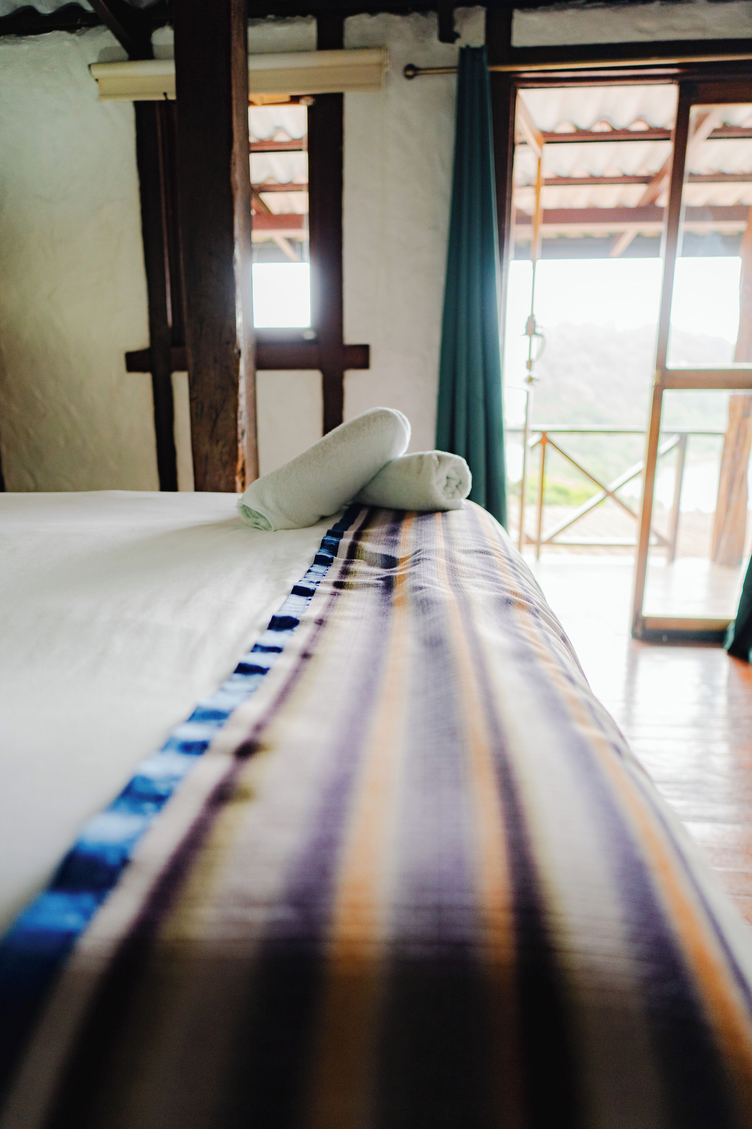 Una cama con una colcha a rayas y toallas enrolladas, un cabecero de madera, y vista a un balcón exterior a través de una ventana.