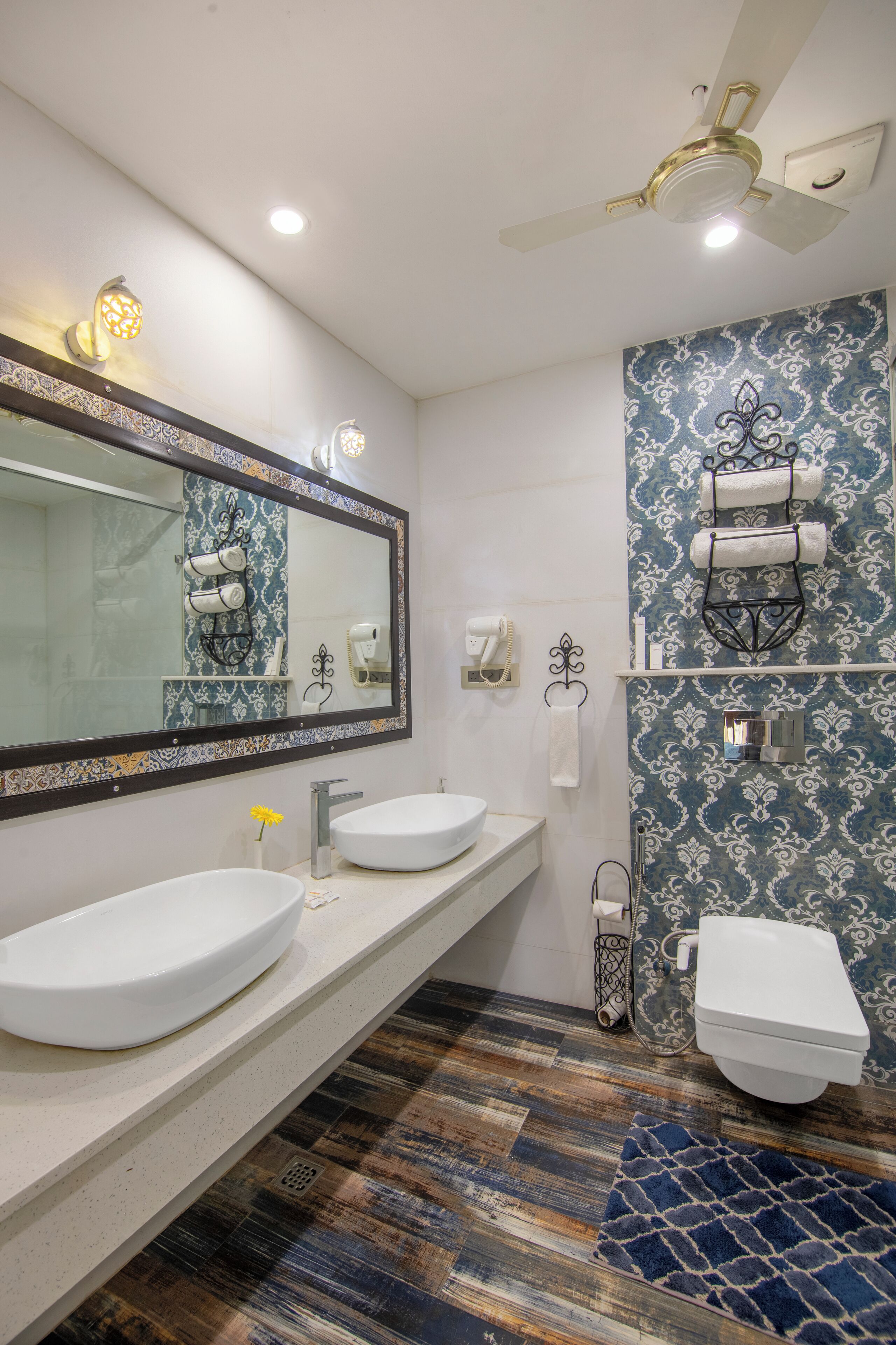 Un baño con dos lavamanos, un inodoro, un espejo, un ventilador de techo y un papel tapiz estampado.