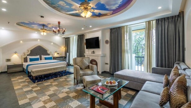 A hotel room with a large bed, a sofa, a glass coffee table, and a ceiling with a cloud mural. - Sterling Marbella Dehradun (Dehradun)