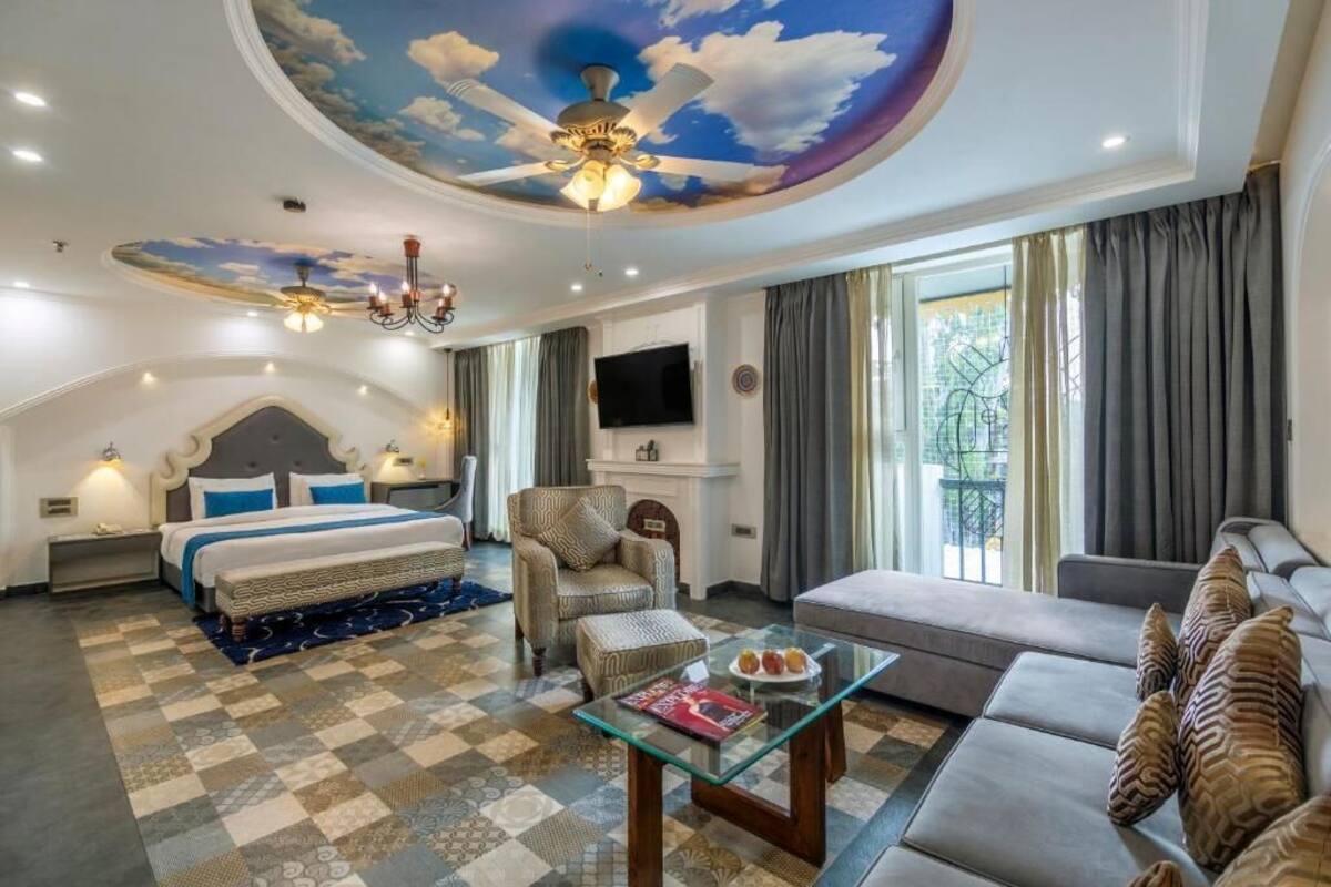 Habitación de hotel con una cama grande, un sofá, una mesa de centro de vidrio y un techo con un mural de nubes.