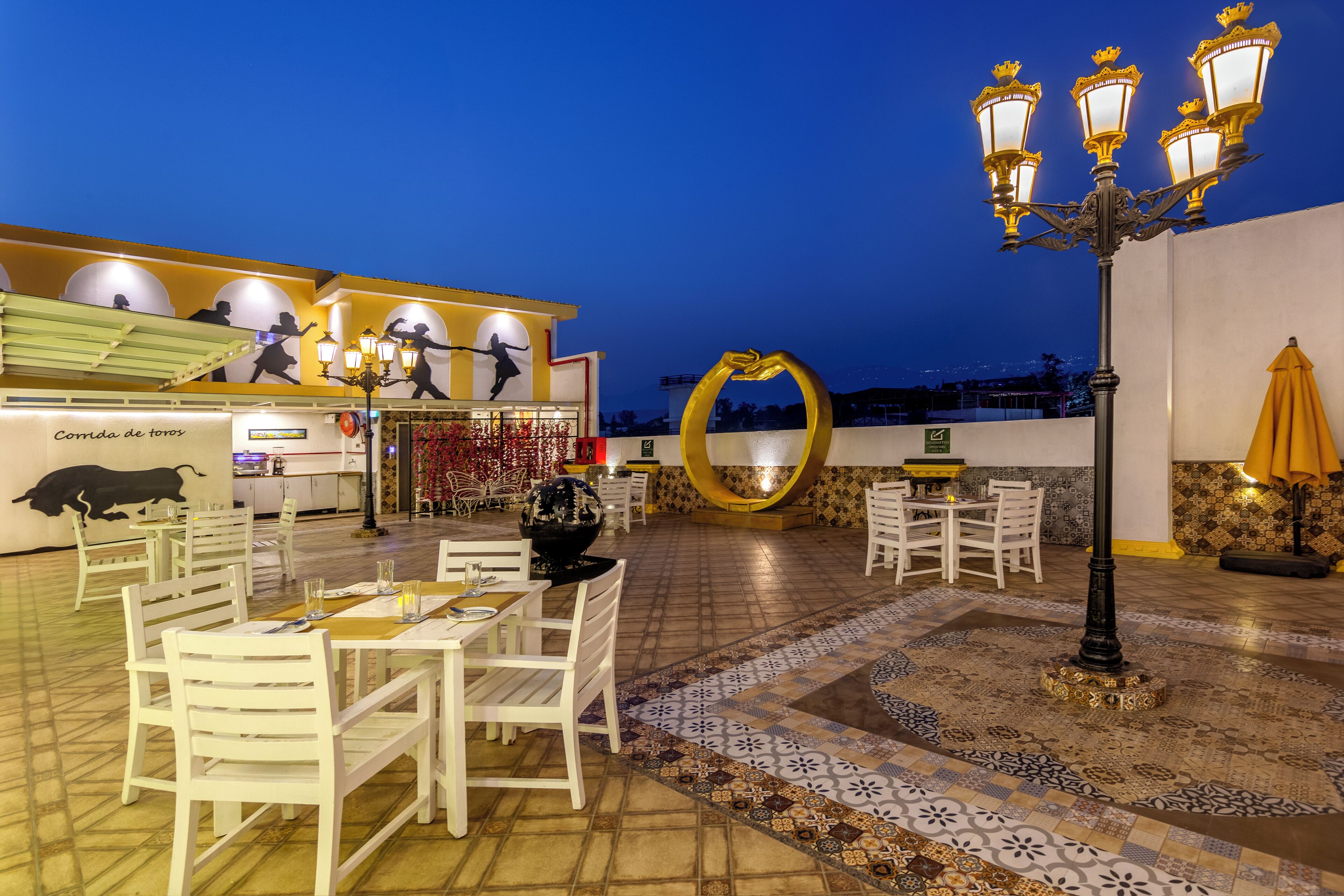Un área de comedor al aire libre con sillas y mesas blancas, una pared decorativa con una escultura de toro y un espejo circular grande.