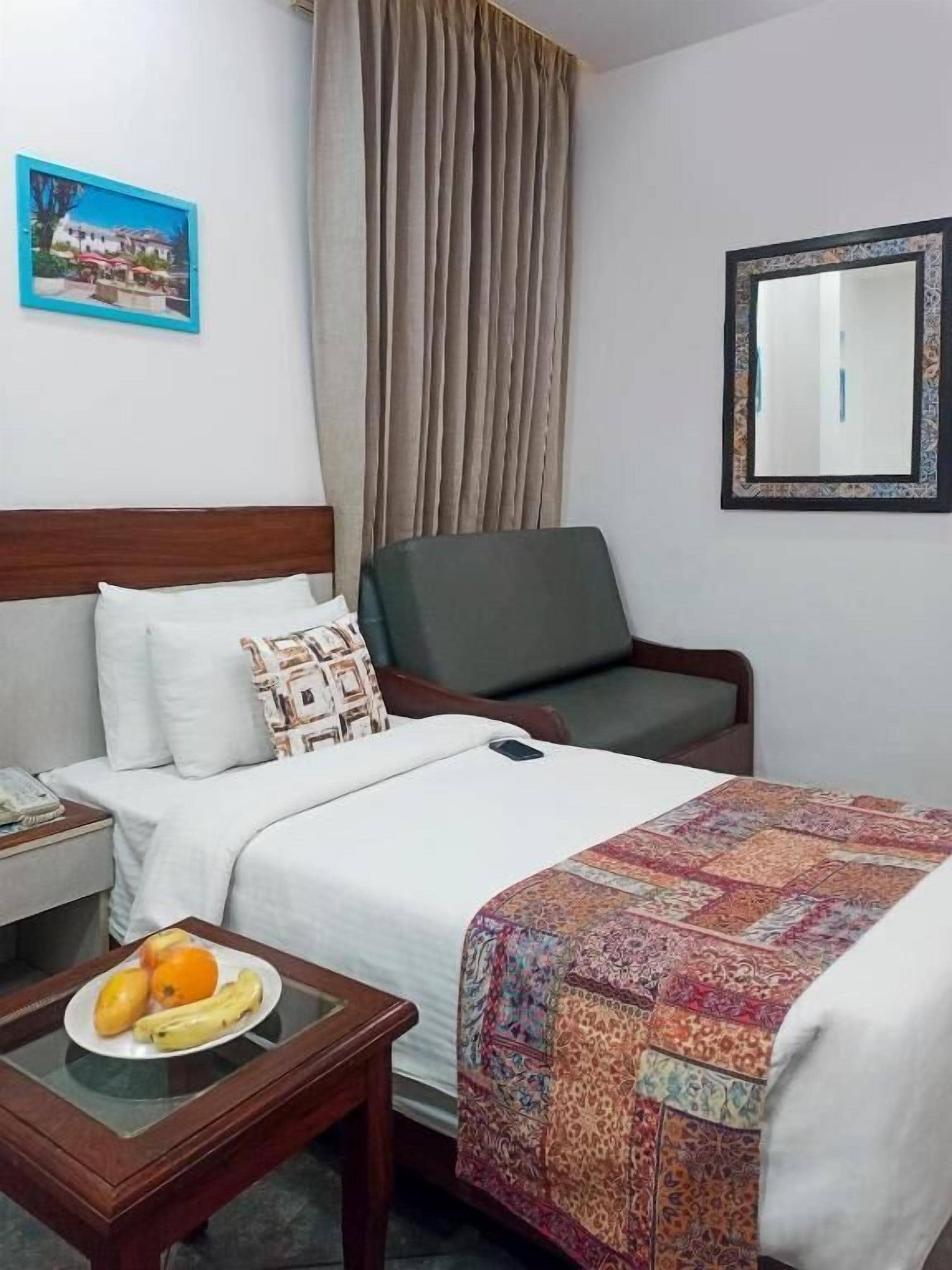 Une chambre d’hôtel avec un lit, une chaise, une table de chevet avec des fruits, un miroir et un tableau au mur.