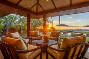 A covered outdoor seating area with chairs and a table, overlooking a scenic view of the ocean and sunset. - Laueki 8 – Stunning Ocean View | Onsite Staff & Daily Housekeeping (Kailua Kona)