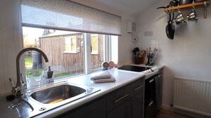A kitchen with a large window, a sink, and a dish rack. There are pots hanging on the wall and a wooden fence visible through the window. - Crossing Cottage Brora (Brora)