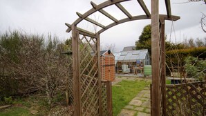 A wooden arbor gate leading to a garden area with a small brick structure and a greenhouse in the background. - Crossing Cottage Brora (Brora)