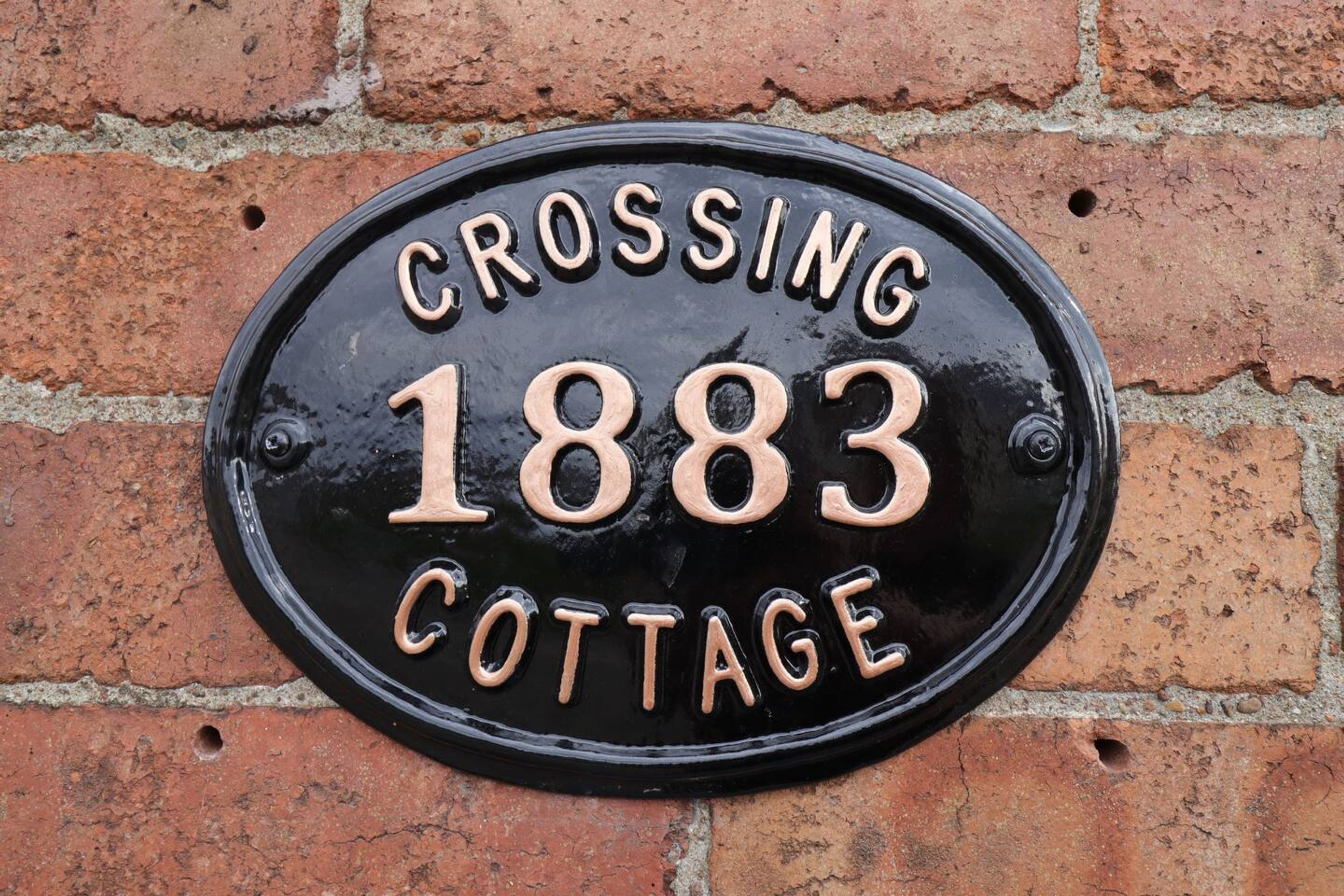 A black metal plaque with the text "Crossing 1883 Cottage" mounted on a brick wall.