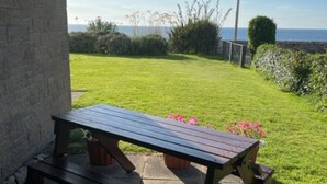 A wooden picnic table with a bench, set on a patio with a view of a grassy area and a body of water in the background. - No 1 The Links Apartment Brora (Brora)