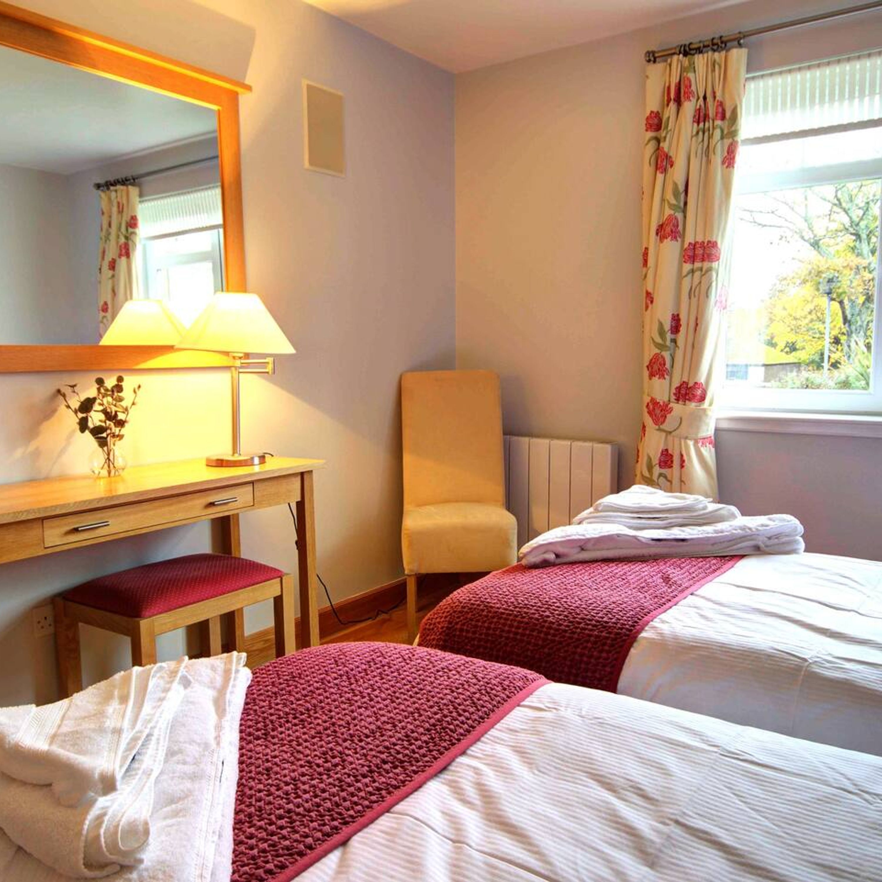 A hotel room with two beds, a desk with a lamp, a chair, and a window with floral curtains.