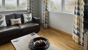 A modern living room with a leather sofa, a coffee table with decorative bowls, and large windows with geometric patterned curtains. - No 2 The Links Apartment Brora (Brora)