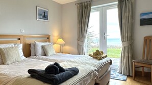 A hotel room with a large bed, a bedside lamp, a view of the sea through the window, and a chair by the window. - No 2 The Links Apartment Brora (Brora)