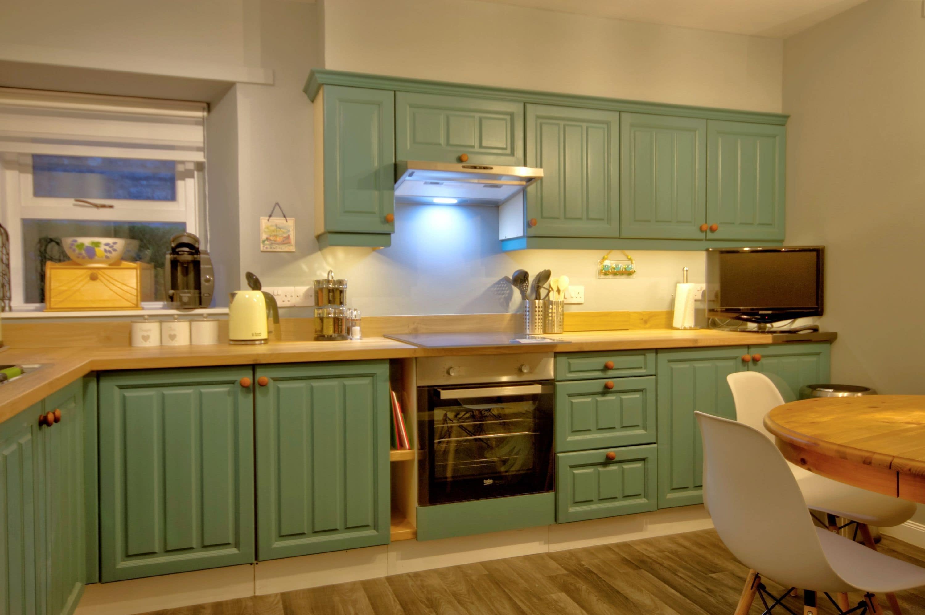 Una cocina con armarios color turquesa, encimera de madera, horno, fregadero y comedor con una mesa redonda y sillas blancas.