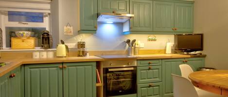 Una cocina con armarios color turquesa, encimera de madera, horno, fregadero y comedor con una mesa redonda y sillas blancas.