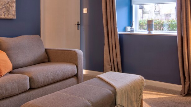 A modern living room with a sofa and ottoman, a window with curtains, and a door. - Otago Cottage Brora (Brora)