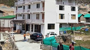 A multi-story building with balconies, a paved area in front, and a mountainous backdrop. - Hotel Pinnacle Peaks (Joshimath)