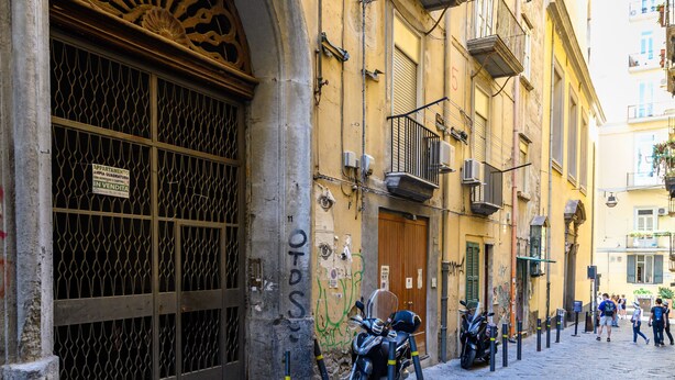 A narrow street with historic buildings, a large arched entrance, and parked motorcycles. - La Stanza del Principe (Naples)