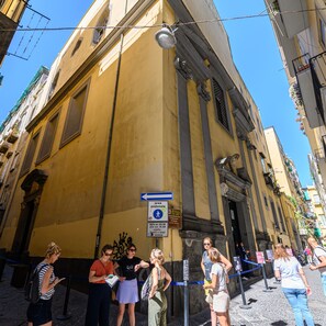 Rua estreita com edifícios amarelos, placa indicando área para pedestres e uma barreira de corda azul.