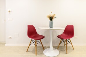 Una habitación con una mesa redonda blanca, dos sillas rojas y un jarrón con plantas secas.