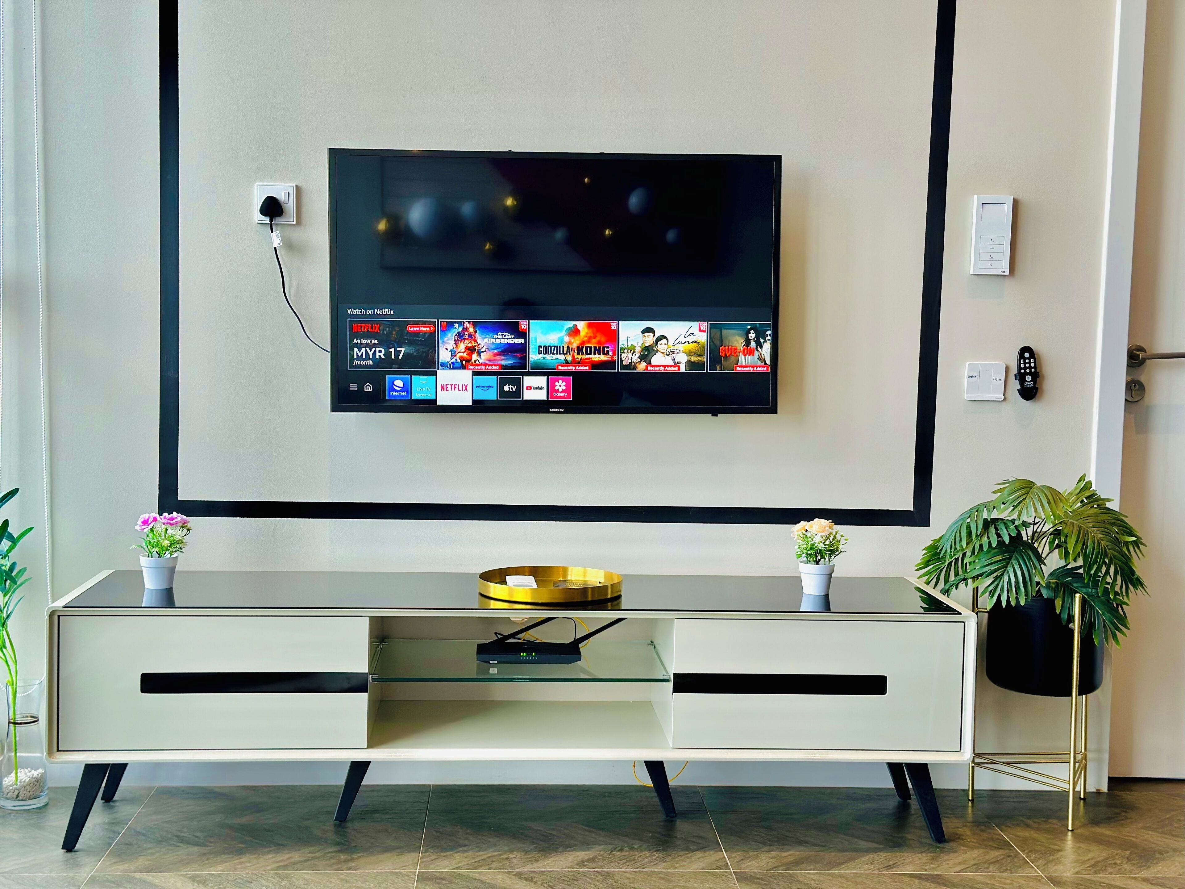 Un salon moderne avec un téléviseur à écran plat fixé au mur, une table basse en verre et deux plantes en pot.