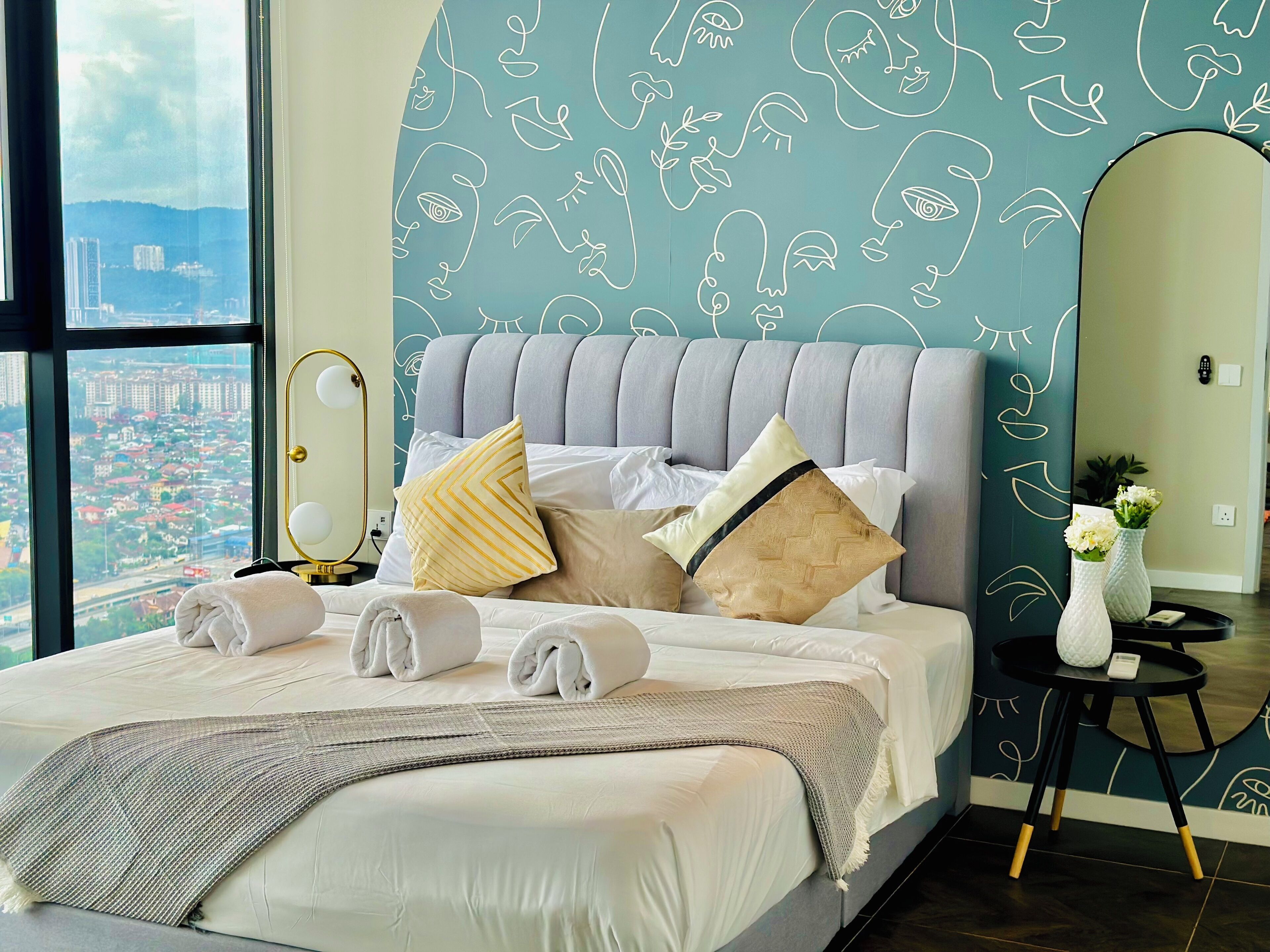 A bed with a view of a cityscape through a large window, a mirror, and a small table with a vase and a remote control.