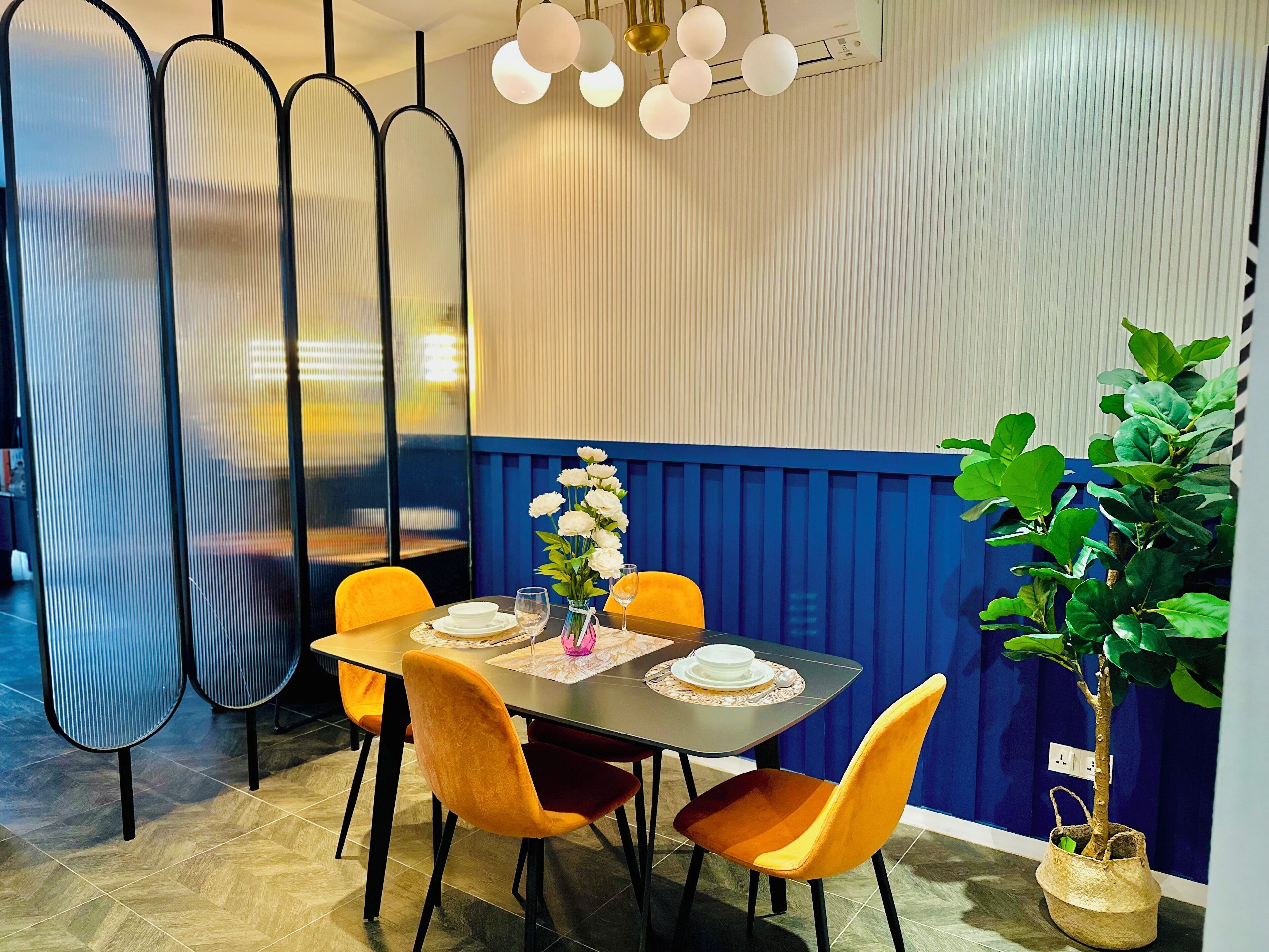A dining area with a table set for four, surrounded by orange chairs, a potted plant, and a decorative wall panel.