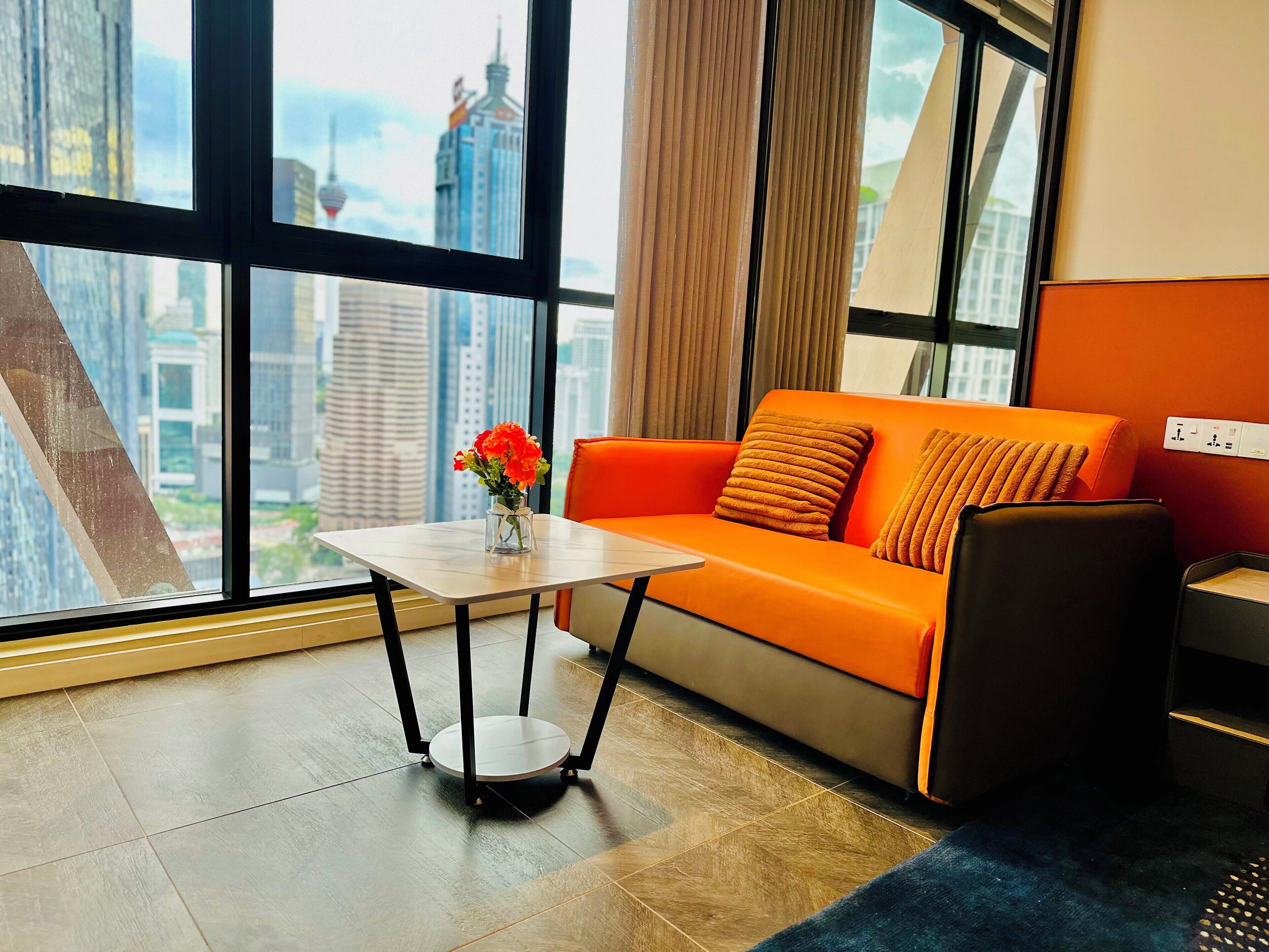 Habitación de hotel moderna con un ventanal grande que ofrece vistas a la ciudad, un sofá naranja, una pequeña mesa con un florero y una silla negra.