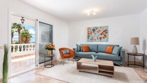 A modern living room with a teal sofa, orange cushions, a wooden coffee table, and a view of palm trees through a glass door. - Casa Orquidea 4 (Adeje Miraverde)
