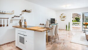 A modern kitchen with a wooden island, white cabinetry, and a dining area with chairs. There is a large window with a view of a mountainous landscape. - Casa Orquidea 4 (Adeje Miraverde)