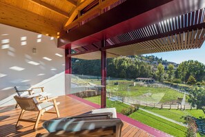 Een modern terras buiten, met houten meubilair en uitzicht op een aangelegde tuin met een hek en bomen.