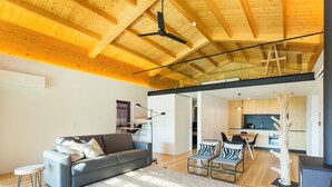 Una sala de estar moderna con techo de madera, un sofá gris, una zona de comedor con sillas y una cocina al fondo.