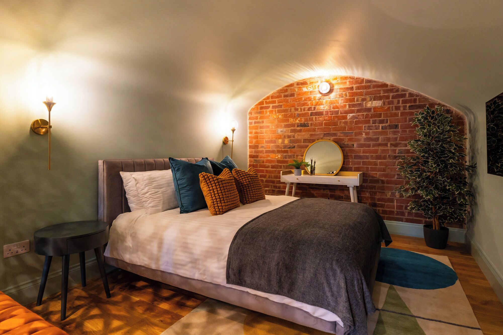 Un dormitorio con arco de pared de ladrillo, una cama con varias almohadas, una mesita de noche con espejo y una pequeña planta.