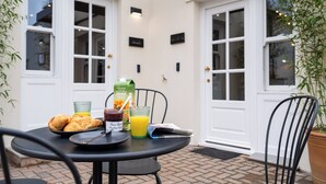 Ein Tisch mit Frühstücksangeboten, darunter Croissants, eine Saftpackung und ein Glas Orangensaft, vor einer weißen Wand mit zwei Türen und einem Blumentopf.