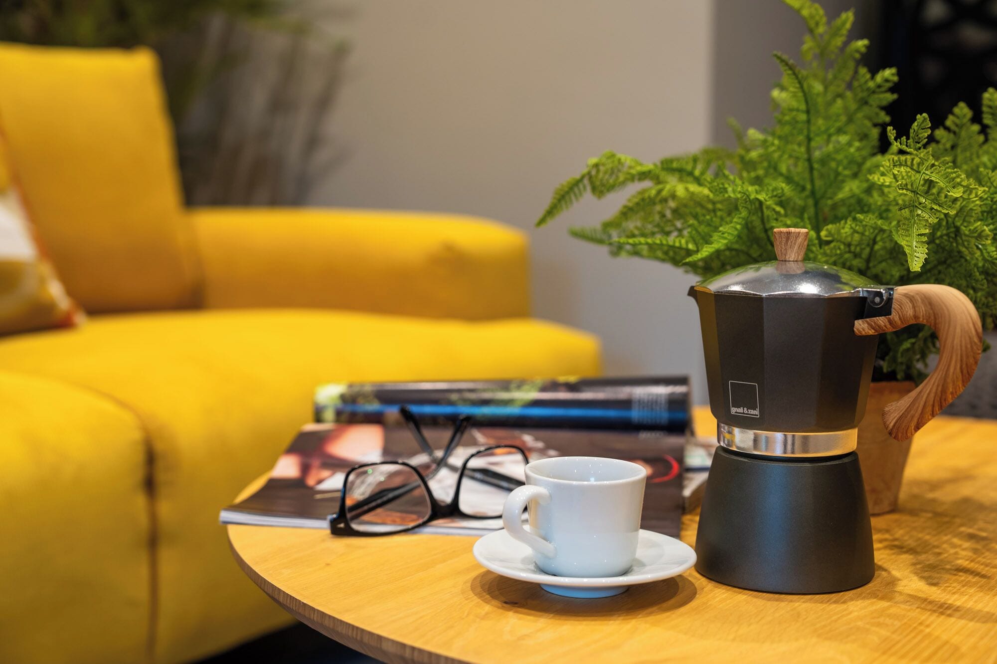 A coffee maker on a wooden table with a white cup and saucer, a pair of glasses, and a magazine.