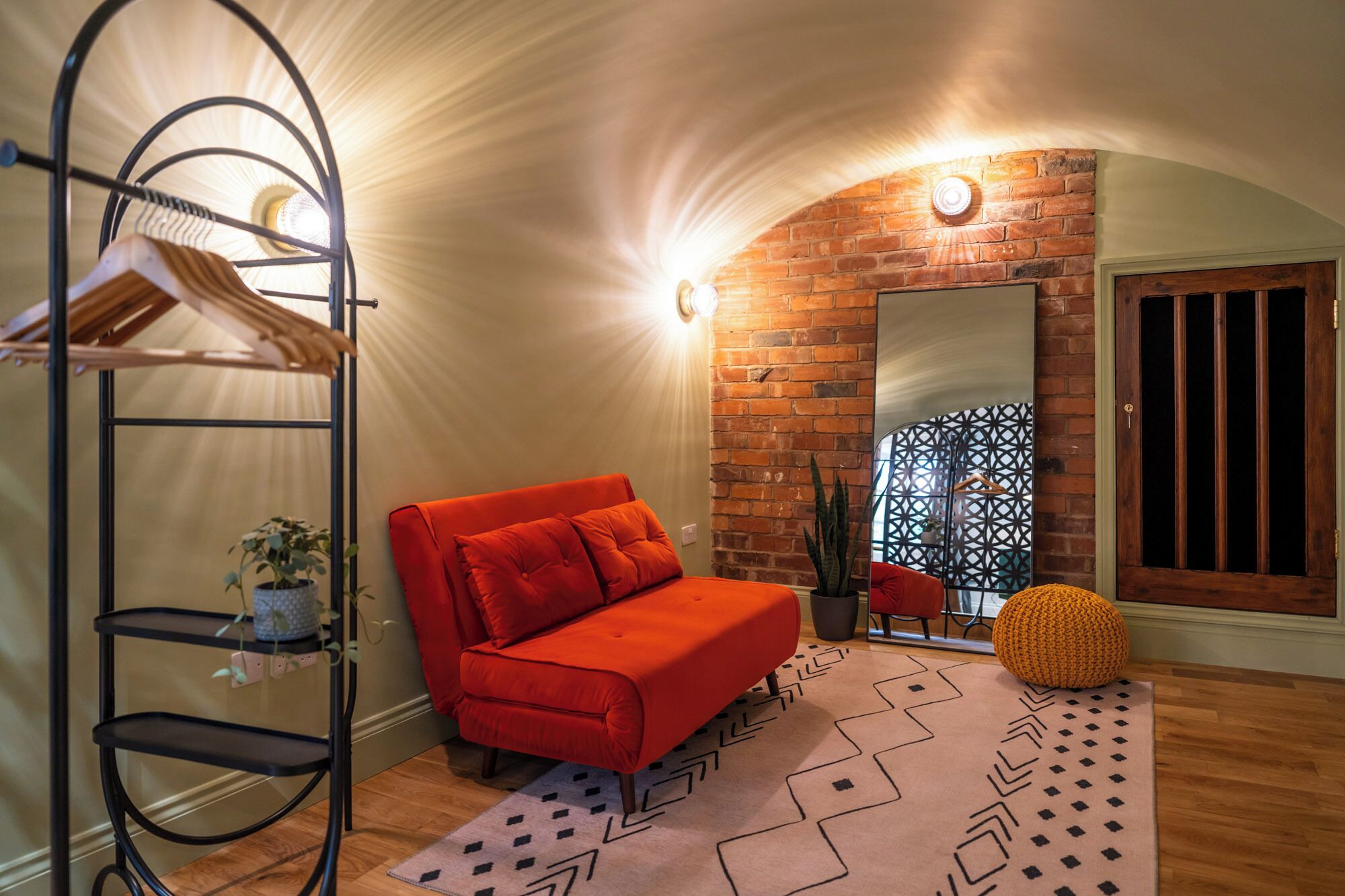 A cozy room with a red sofa, a black metal shelf with clothes, a patterned rug, and a brick wall with a mirror.