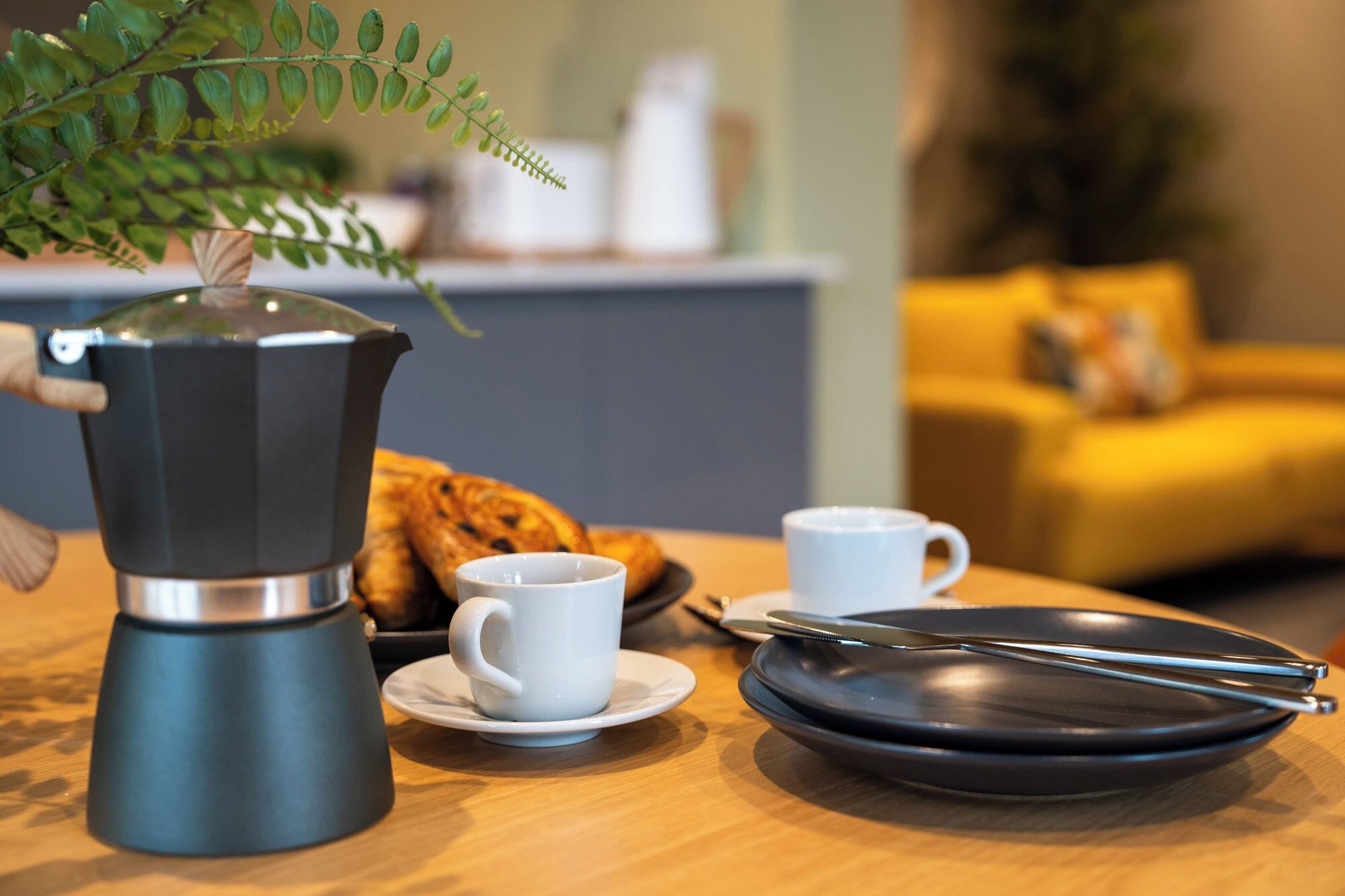 A kitchen counter with a coffee maker, two cups, a plate, and a pair of tongs.