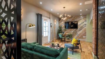 A modern living room with a green sofa, a dining area with a table and chairs, a floor lamp, and a TV mounted on a marble wall.