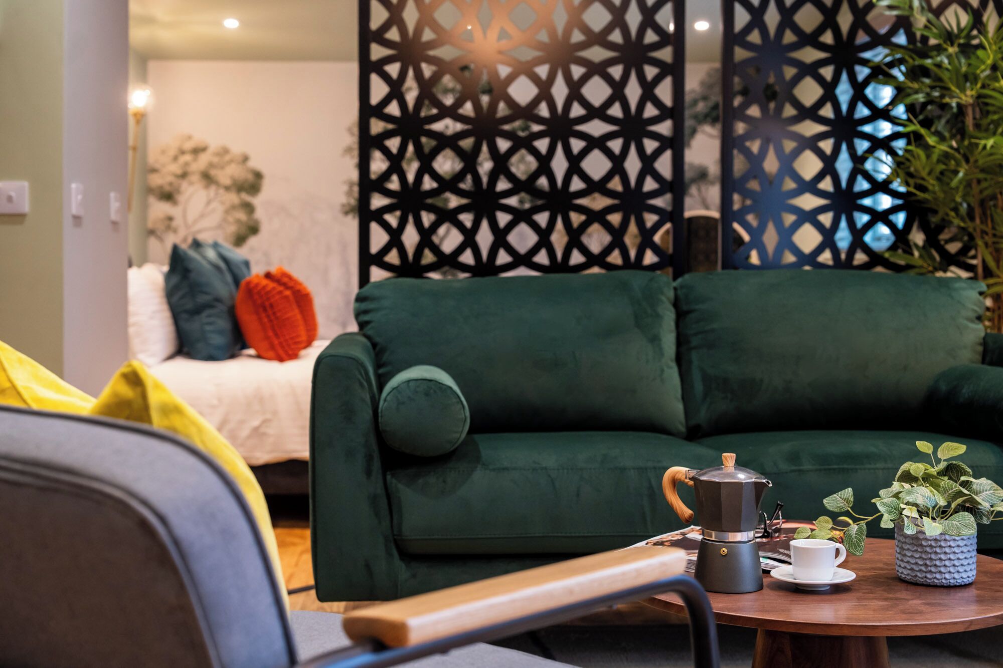 Un salon accueillant avec un canapé vert, une table basse sur laquelle se trouvent une cafetière et une tasse, et un élément de séparation décoratif orné de motifs géométriques.