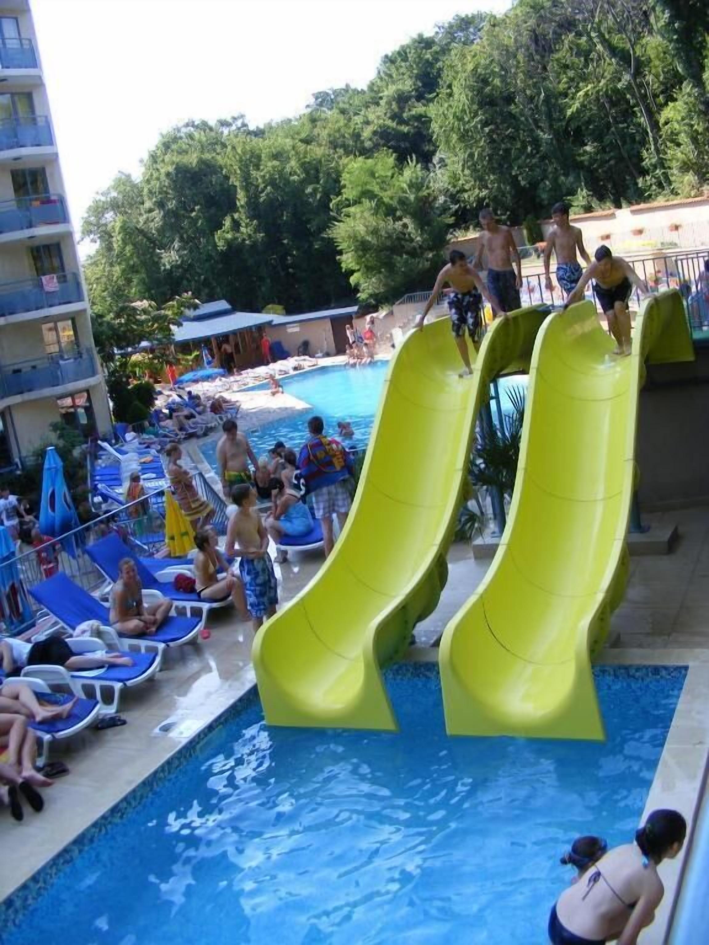 A pool area with two yellow slides, lounge chairs, and a building in the background.