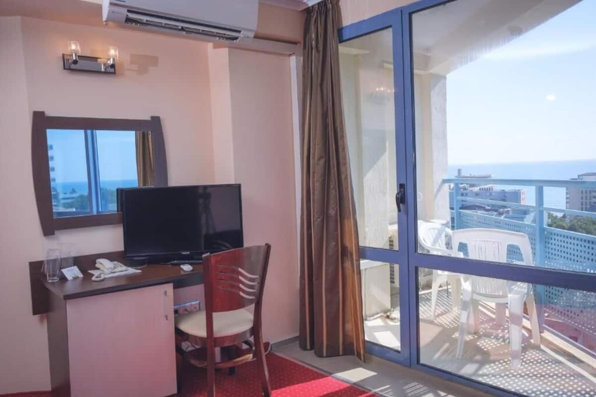 A hotel room with a desk, TV, and a view of the city through a large sliding glass door.