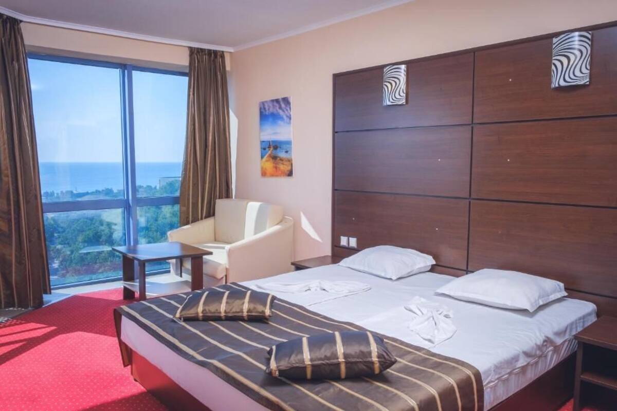 A hotel room with a large bed, a chair, a small table, and a view of the sea.