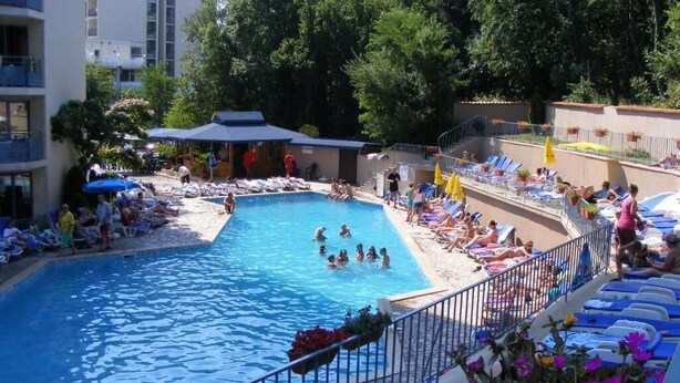 A swimming pool with lounge chairs and umbrellas, surrounded by buildings and trees. - Regina Hotel (Varna)