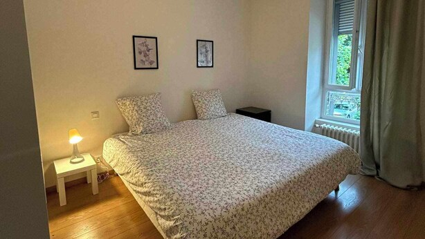 A bedroom with a bed, a nightstand, a lamp, two framed pictures, a window with curtains, and a ceiling light fixture. - Maison Familiale au Centre de Guingamp Animaux de Compagnies Acceptés (Guingamp)
