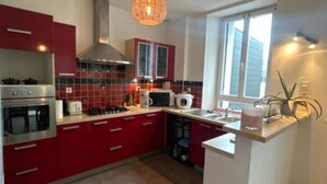 A modern kitchen with red cabinets, stainless steel appliances, a large window, and a pendant light. - Maison Familiale au Centre de Guingamp Animaux de Compagnies Acceptés (Guingamp)