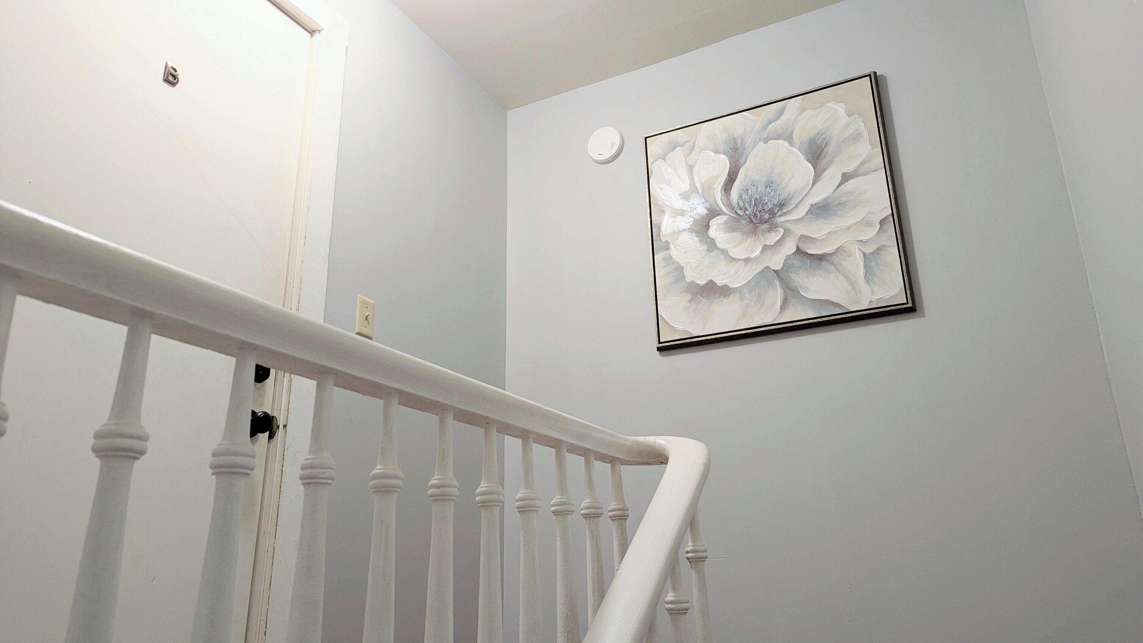 A staircase with white balusters and a white handrail, leading to a room with a light gray wall and a framed floral artwork.