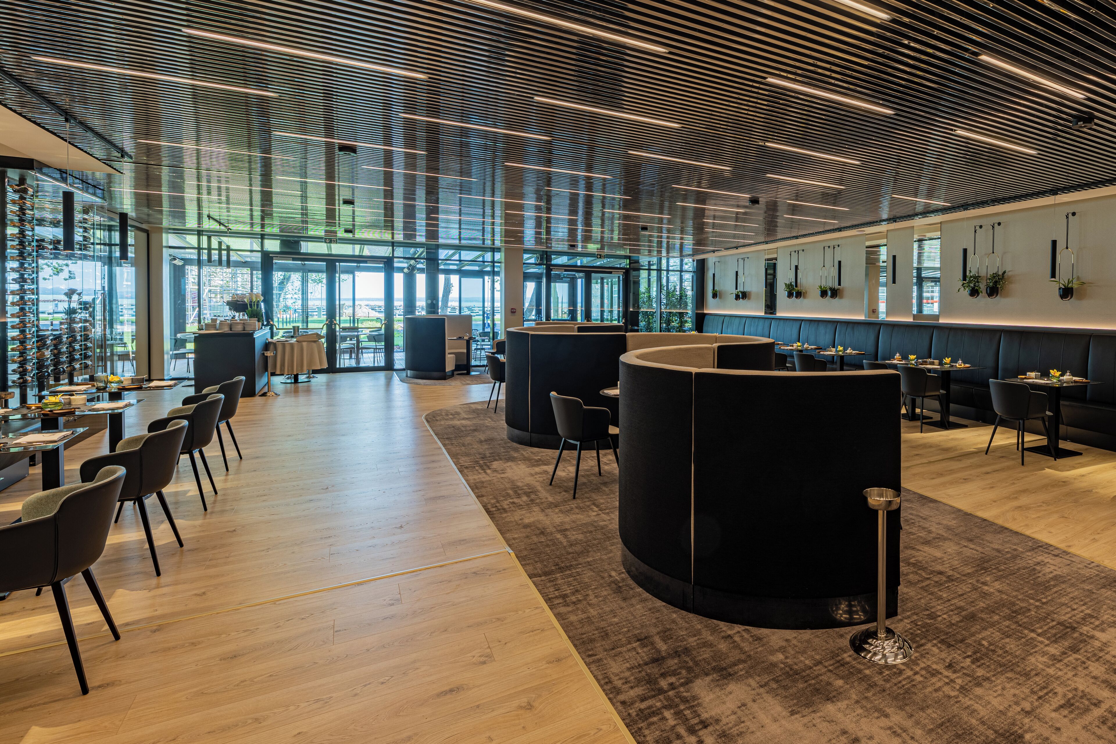 A modern interior with a wooden floor, black seating, and a glass wall offering a view of the outdoors.